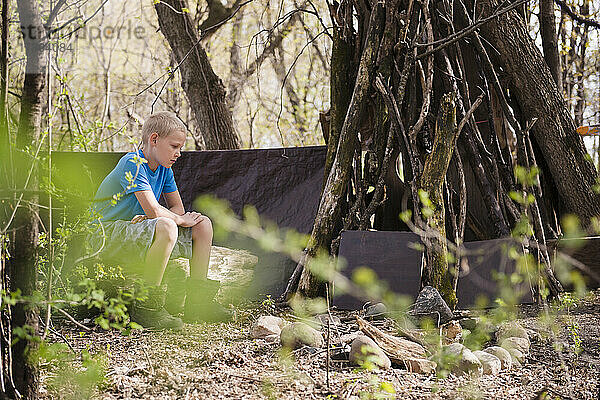 Junger blonder Junge sitzt bei seiner Festung im Wald.
