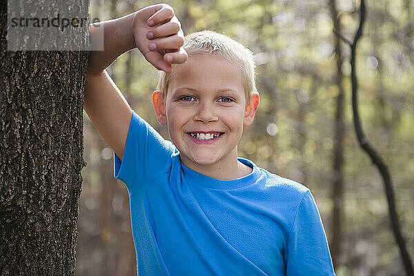 Fröhlicher blonder Junge  der sich an einen Baum im Wald lehnt.
