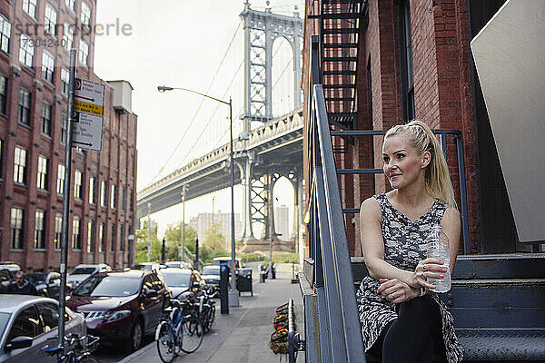 Positive Frau  die auf einer Gebäudetreppe vor einer majestätischen Hängebrücke sitzt