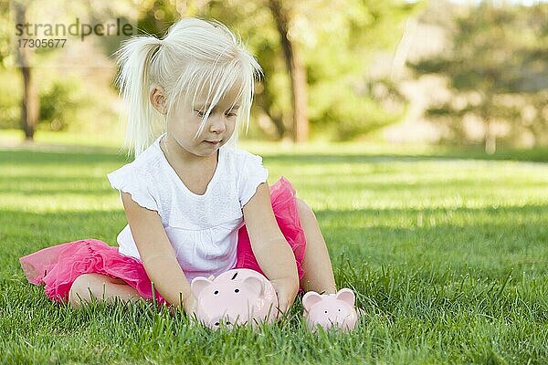 Nettes kleines Mädchen mit Spaß mit ihren großen und kleinen Sparschweinen draußen auf dem Gras