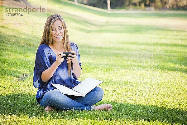 Lächelnde junge Frau mit Buch und Handy im Freien im Park