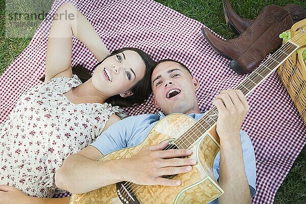 Glückliches gemischtrassiges Paar auf Picknickdecke im Park spielt Gitarre und singt Lieder
