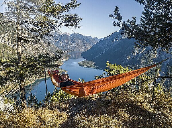Mann fotografiert sich mit Handy  Wanderer sitzt in Hängematte mit Ausblick auf Berge mit See  Plansee  Ammergauer Alpen  Bezirk Reutte  Tirol  Österreich  Europa