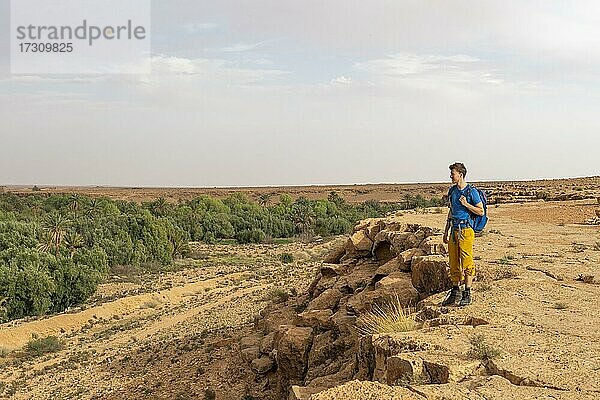 Junger Mann an einer Felsabbruchkante  hinten Tal voller Palmen an der Oase Source Bleu  Blaue Quelle  Madkhal Meski  Marokko  Afrika