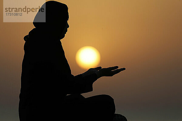 Silhouette einer muslimischen Frau in Abaya  die bei Sonnenuntergang mit ihren Händen betet  Vereinigte Arabische Emirate  Naher Osten