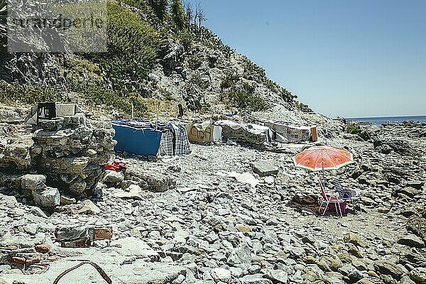 Zelte und selbsterrichtete Hütten an der Steilküste am Küstenabschnitt El Sarchal  an der Steilküste haben mehr als 200 Migranten temporär ihr Lager aufgeschlagen  Küste teilweise schwer zugänglich und teilweise von der Küstenstraße nicht einsehbar  rechts Strandstühle und Sommerschirm von Badegästen  Ceuta  Spanien  Europa