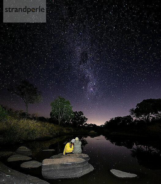 Mand und Frau sitzen zusammen auf einem Felsen neben einem Fluss und beobachten die Milchstraße