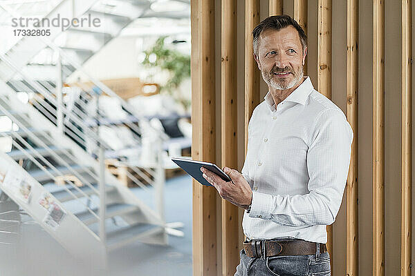 Nachdenklicher Geschäftsmann  der ein digitales Tablet hält  während er an der Wand im Büro steht