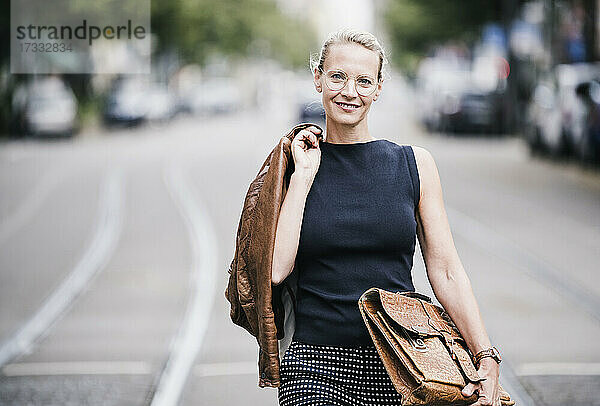 Lächelnde Geschäftsfrau mit Jacke und Tasche beim Gehen auf der Straße