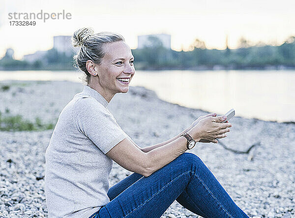 Glückliche reife Frau mit Handy in der Hand am Flussufer sitzend
