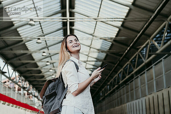 Fröhliche junge Passagierin mit Rucksack  die am Bahnhof wegschaut