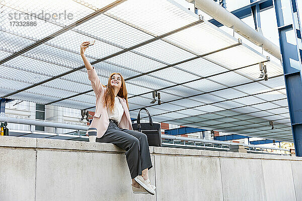 Lächelnde Geschäftsfrau  die ein Selfie mit ihrem Mobiltelefon macht  während sie auf einer Stützmauer am Bahnhof sitzt