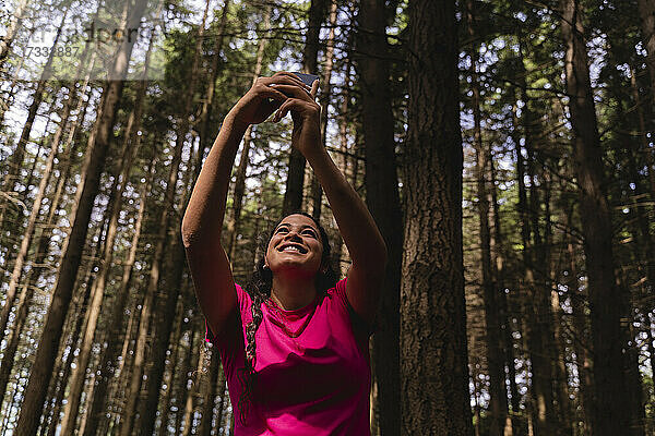 Lächelnde weibliche Wanderin nimmt Selfie durch Handy mit Bäumen im Hintergrund im Wald