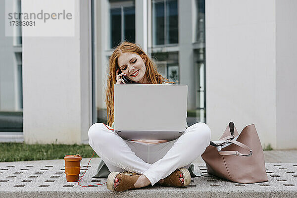 Geschäftsfrau sitzt im Schneidersitz auf einer Bank und spricht mit einem Smartphone  während sie einen Laptop benutzt
