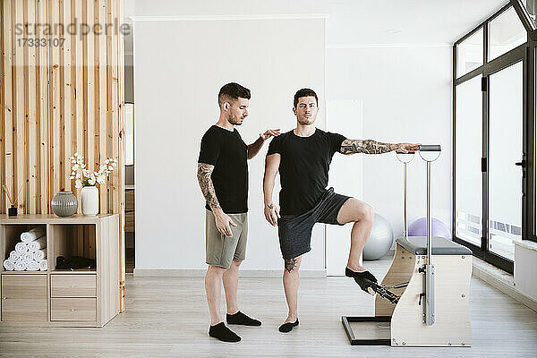 Mann trainiert auf einem Stabilitätsstuhl  während ein männlicher Ausbilder im Pilates-Studio unterstützt