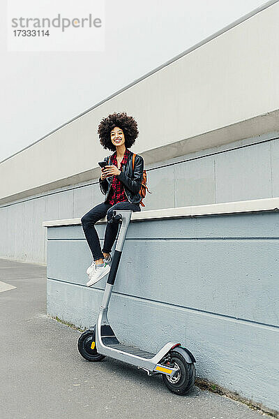 Junge Frau mit Mobiltelefon auf Stützmauer sitzend mit Elektro-Scooter