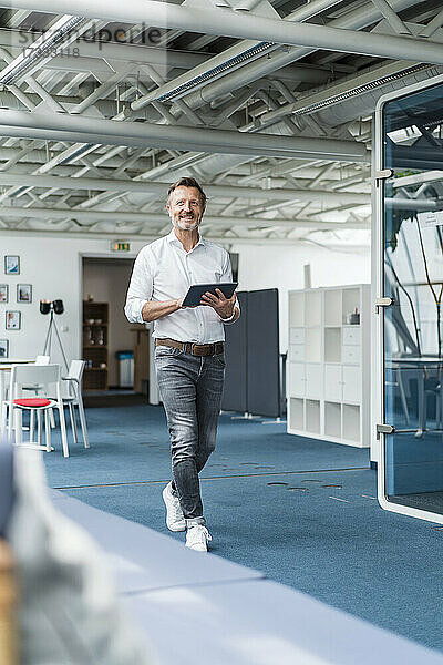 Lächelnder Geschäftsmann  der ein digitales Tablet hält  während er im Büro spazieren geht
