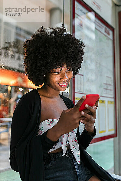 Afro-Frau lächelt bei der Benutzung eines Smartphones an der Bushaltestelle