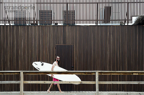 Lächelnder Mann  der ein Surfbrett trägt  während er an einer Mauer vorbeiläuft