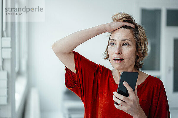 Geschäftsfrau mit offenem Mund und Hand in den Haaren hält Smartphone im Büro