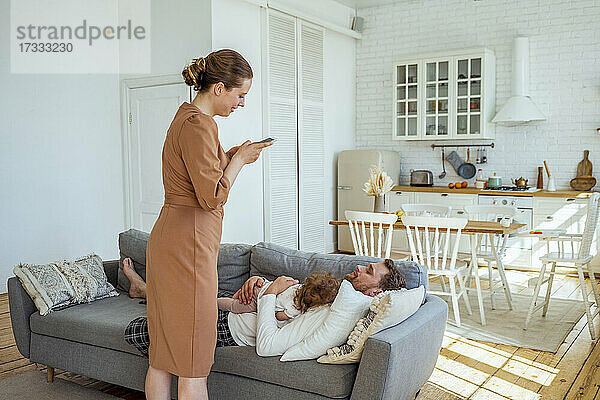 Frau fotografiert Mann  der mit seiner Tochter auf dem Sofa zu Hause schläft