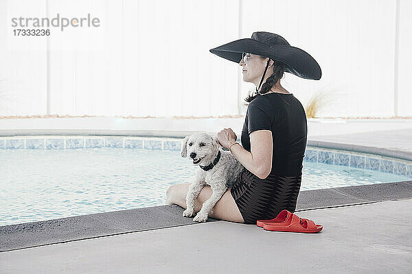 Frau mit Hut schaut weg  während sie mit ihrem Hund am Pool im Innenhof sitzt