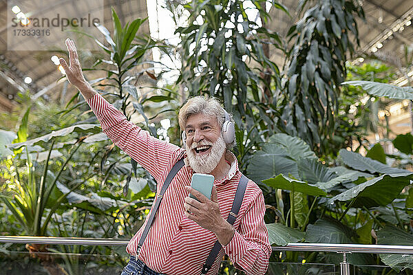 Fröhlicher reifer Mann mit erhobener Hand tanzt beim Hören von Musik vor Pflanzen