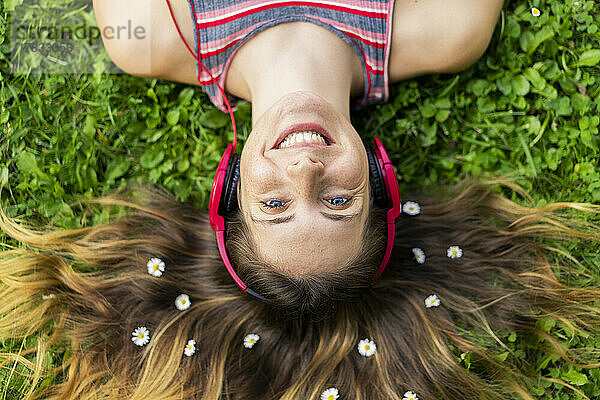 Glückliche junge Frau mit Blumen im Haar  die im Gras liegt und über Kopfhörer Musik hört
