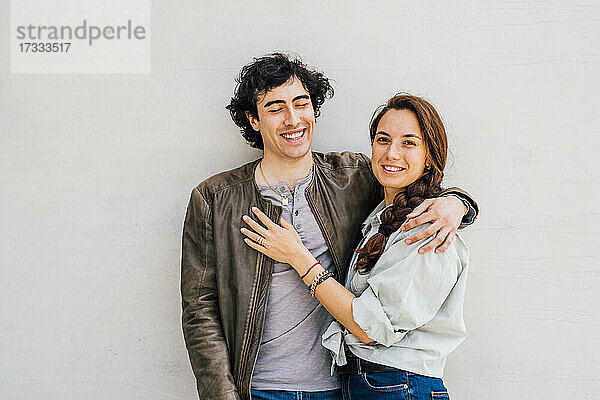 Lächelndes Paar mit umschlungenen Armen vor einer Wand stehend