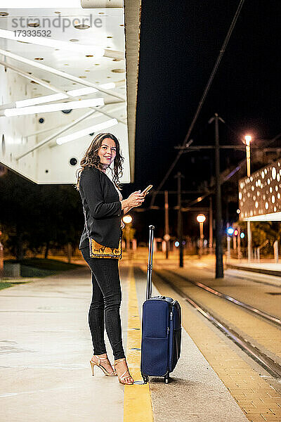 Lächelnde weibliche Fachkraft mit Gepäck  die ihr Handy in der Hand hält  während sie auf eine nächtliche Straßenbahnstation wartet