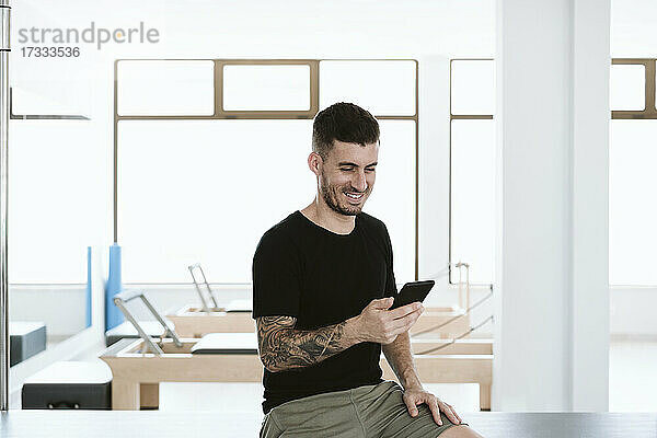 Lächelnder männlicher Besitzer  der ein Smartphone benutzt  während er im Pilates-Studio sitzt