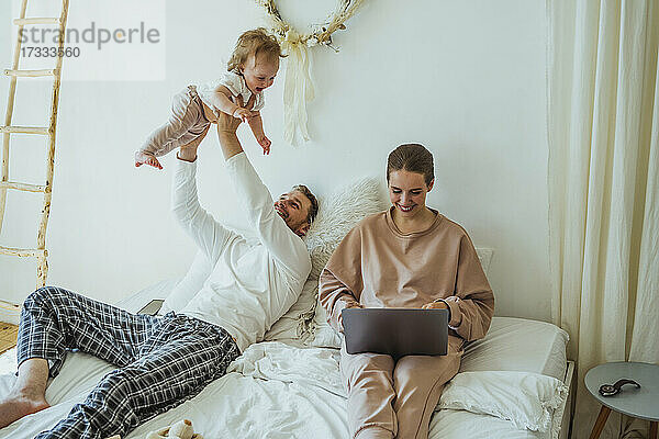 Lächelnde Frau mit Laptop von spielerischen Mann hebt Tochter auf dem Bett zu Hause