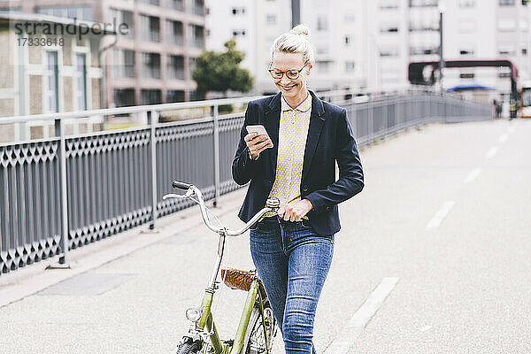 Blonde Pendlerin  die auf einer Brücke Fahrrad fährt und dabei ein Mobiltelefon benutzt