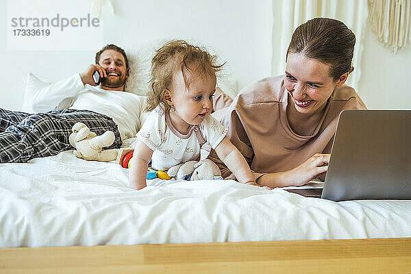 Lächelnde Frau mit Laptop  die ihre Tochter anschaut  während der Mann im Schlafzimmer mit dem Handy telefoniert