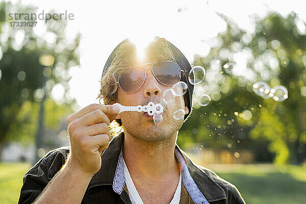 Junger Mann mit Sonnenbrille beim Blasen von Seifenblasen im Park