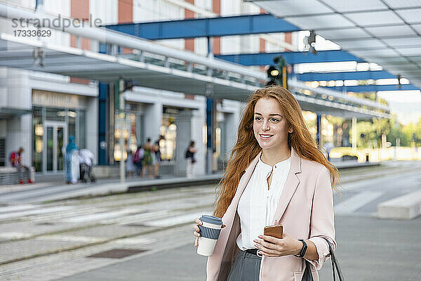 Lächelnde Geschäftsfrau mit Smartphone  die am Bahnhof wegschaut
