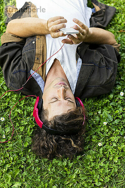 Junger Mann  der sein Smartphone benutzt und über Kopfhörer Musik hört  während er im Gras liegt