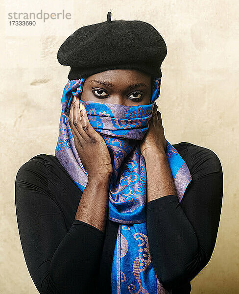 Junge Frau bedeckt ihr Gesicht mit einem Kopftuch vor einer beigen Wand