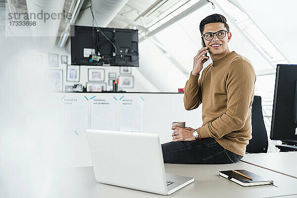 Lächelnder Geschäftsmann  der mit seinem Handy telefoniert  während er im Büro am Laptop sitzt