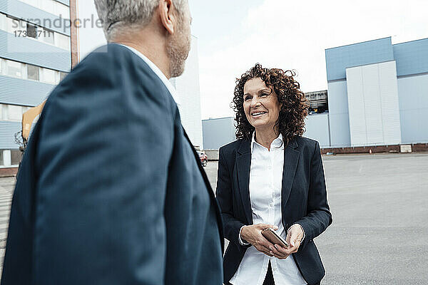 Lächelnde Geschäftsfrau im Gespräch mit einem Kollegen  während sie auf dem Fußweg steht