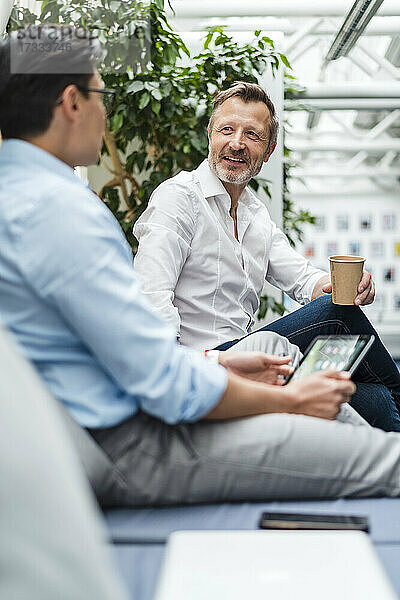 Lächelnder Geschäftsmann mit Kaffeetasse  der neben einem Kollegen im Büro sitzt