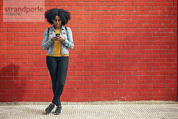 Junge Frau benutzt ihr Smartphone  während sie vor einer roten Backsteinmauer steht