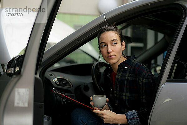 Mechanikerin mit Kaffeetasse  die wegschaut  während sie im Auto sitzt