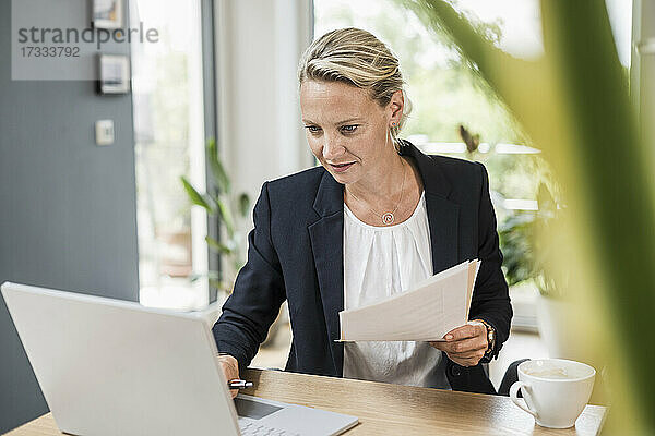 Geschäftsfrau hält ein Dokument  während sie einen Laptop im Büro benutzt