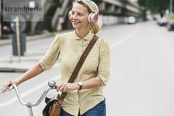 Blonde Geschäftsfrau mit Kopfhörern lächelt  während sie auf der Straße Fahrrad fährt