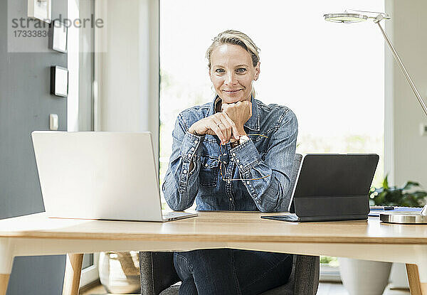 Lächelnde Freiberuflerin mit Laptop und digitalem Tablet  die mit der Hand am Kinn im Home Office sitzt