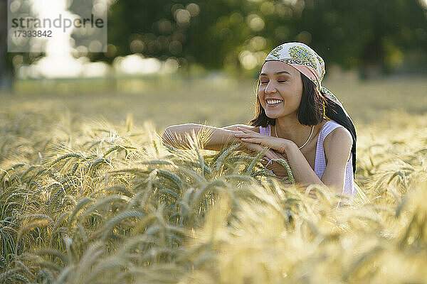Lächelnde junge Frau steht mit geschlossenen Augen und genießt in einem Weizenfeld