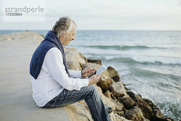 Glücklicher reifer Mann mit digitalem Tablet am Meer sitzend