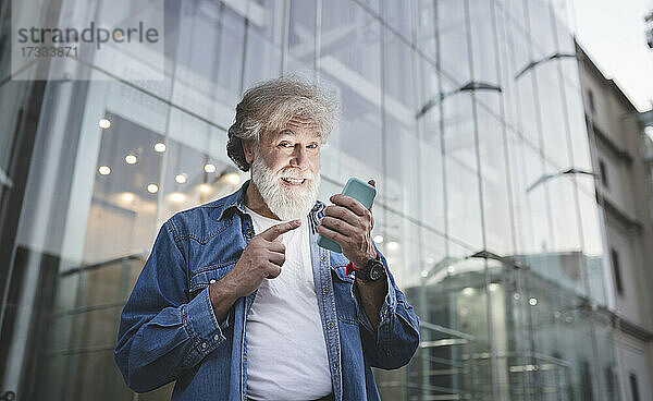 Glücklicher reifer Mann  der auf sein Smartphone vor einem Glasgebäude zeigt