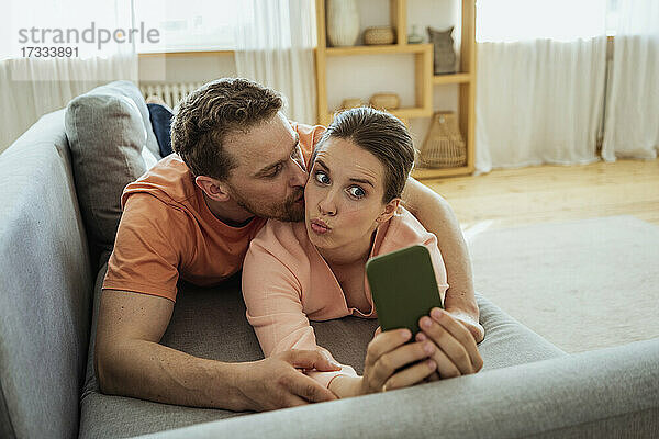 Mann küsst Frau  die ein Selfie mit ihrem Handy macht  während sie zu Hause auf dem Sofa liegt